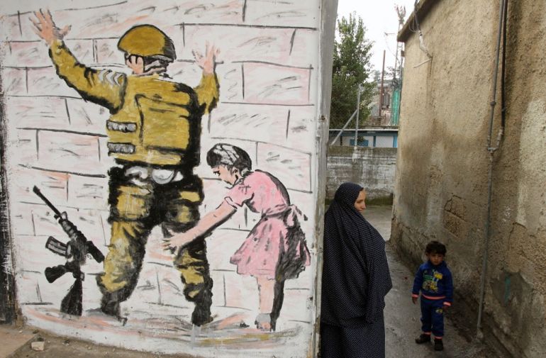 A Palestinian woman stands with a child in an alley next to reproduction of a mural by British street artist Banksy, originally painted on the wall of the West Bank in Bethlehem, at al-Aroub Palestinian refugee camp, north the West Bank town of Hebron on February 11, 2016. [Hazem Bader/AFP]
