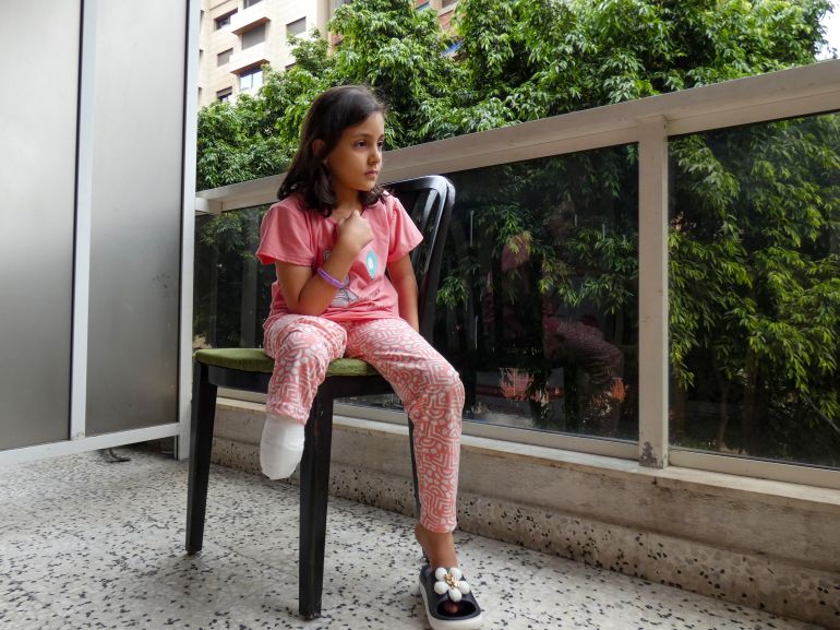 A little girl in pink pajamas sits on a plastic chair in a balcony, one plastic slipper with a flower on her left foot. Her right leg is amputated, a stump hangs below her knee.