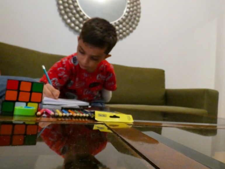 A little boy in a red t-shirt leans over, drawing with his right hand. His left arm is a stump, tucked by his side. In front of him are some coloured pencils and a Rubiks Cube
