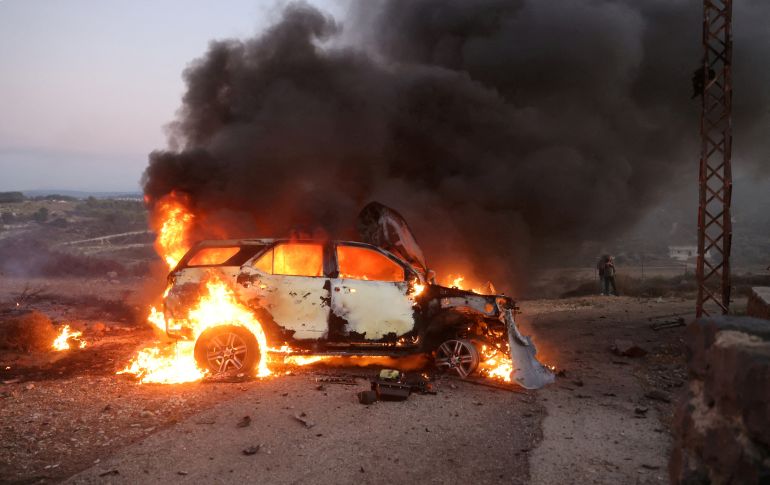Flames burn brightly within the charred shell of a small sedan car, with black smoke billowing out of it.