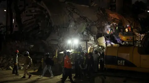 Reuters A team of emergency workers using heavy machinery to search a collapsed building, lit by bright headlights in darkness