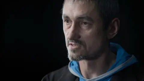 Portrait image of Oleksii Sivak, who has a short beard and moustache, looking to the left of the camera, wearing a light blue hoodie under a black jacket 