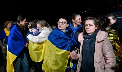 Dmytro Smolyenko / Ukrinform/Future Publishing via Getty Images Several women wrapped in blue and yellow Ukrainian flags. Two women are hugging the background, others are smiling. Liudmyla is looking to her right while holding the hand of a woman in a pink coat on her left.