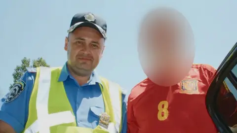 OK.ru Temerbek is pictured in a black police cap, blue police shirt and yellow hi-vis vest, looking at the camera with a slight smile. Next to him is a figure in a red football shirt, whose face has been blurred.
