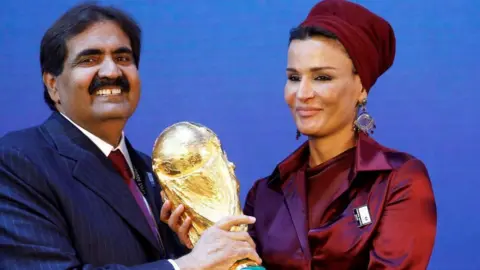 Reuters Sheikh Hamad grins as he grips the trophy with his wife. He is dressed in a blue pin strip suit and his wife is in a maroon coloured outfit.