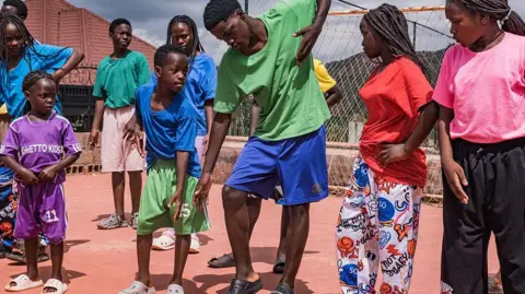 AFP via Getty Images Members of the Ugandan dance group the Ghetto Kids rehearse at their home in Nakawuka, Uganda, on 25 May 2026. They are wearing an array of brightly coloured T-shirts and mimicking the moves of an adult man choreographer. 