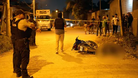 Lee Durant/BBC Police stand beside a corpse, lying next to a toppled motorcycle on the street. Onlookers watch from behind police tape. 