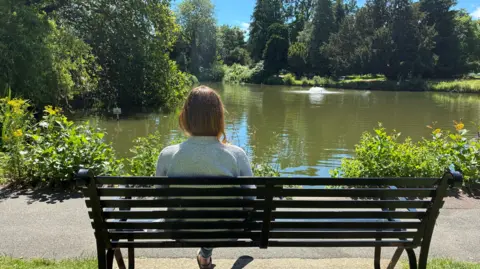 A woman is sat on a bench next to a lake. You can see the back of her body as she looks at the water. There are green plants around the water's edge, trees in the background and a fountain in the middle of the lake.