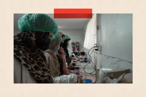 A row of women in headscarves and hair nets work at a long table with jam jars on it
