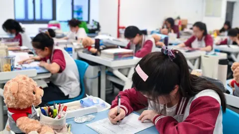 Getty Images A teenager sits at a desk with her head down looking at a paper. She is writing on it with black pen. Stationery and a teddy bear sits on her desk. In the background are other teenagers at desks, all spaced out, in a classroom.