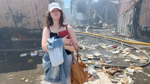 BBC A young woman clutches a book to her chest and looks sad. Behind her is a bombed-out market structure. Destroyed books litter the ground around her