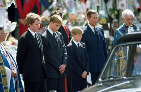 Jeff Overs/BBC Earl Spencer with Princes William and Harry, and Prince Charles at the funeral of Diana, Princess of Wales, standing alongside members of the clergy to the side of the funeral procession, with floral tributes faintly visible in the background