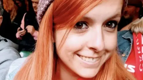 Cornish Family Danielle is shown smiling and wearing a beanie hat in a crowd. Her hair is ginger coloured and she is looking at the camera with her brown eyes.