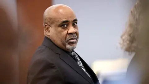 STEVE MARCUS / POOL / AFP via Getty Images A man wears a black suit and patterned tie. He has a white goatee and serious expression.
