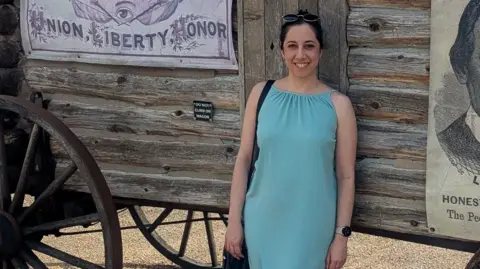 Elena Chrysostomou Elena Chrysostomou wearing a long light blue dress, standing smiling in front of what appears to be a wooden wagon, with a window covering with the words Union, Liberty, Honor on it