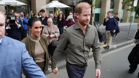 PA Media Prince Harry, wearing a khaki shirt and navy trousers, holds Meghan's hand and as they cross a road, protected by a man in a blazer clearing the way for them to walk. In the background are a crowd of people taking photos.