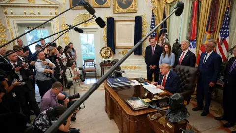 Getty Images Reporters in the Oval Office with boom mics up to Trump and other officials standing behind him at his desk