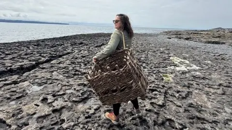 Mary Timby Brunette Colorado native Mary Timby, 39,  wears sunglasses and tennis shoes as she holds a woven basket over her shoulder on an Irish shoreline, the sea and broody skies visible in the background