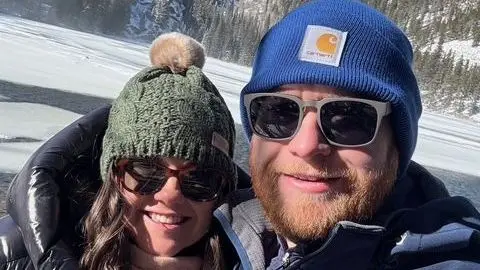 Mary Timby Timby and Mangan both wear winter knit caps, sunglasses and heavy coats in the Colorado snow, a forest treeline visible behind them