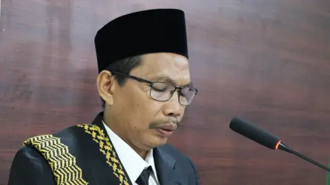 Abu Barokat Facebook Abah wears a black kopi on his head, and a black suit with black tie and white shirts, with a black and yellow sash draped over one shoulder. He is speaking at a podium, a microphone in front of him. 