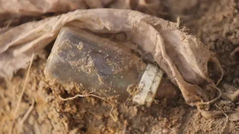 Kumanan Kanapathippillai, BBC A baby's bottle is partially covered in dirt at the grave site.