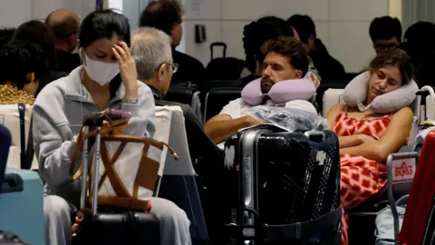 Reuters Several people sat among bags in an airport departure lounge. A woman with a medical face mask on looks down at a smart phone, a man and woman are asleep behind her. 