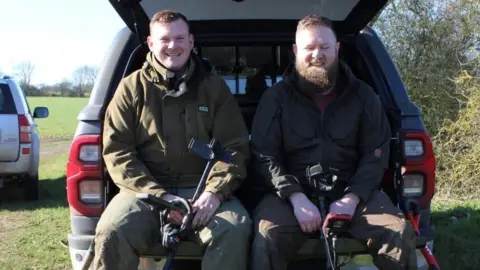 Aaron Pizzey Two men sitting on the edge of the open boot of a dark SUV-type vehicle parked in a field. They are both smiling, wearing dark long-sleeved jackets and dark trousers and have short brown hair. The man on the right has a brown beard. 