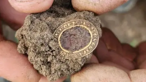 Aaron Pizzey A gold oval with a dark centre. It is embedded in a clod of soil and being held between a man's fingers. 