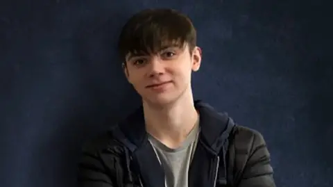 Tanya Whitaker/JustGiving Samuel Whitaker seen on a blue backdrop wearing a grey top, dark hoodie and jacket.