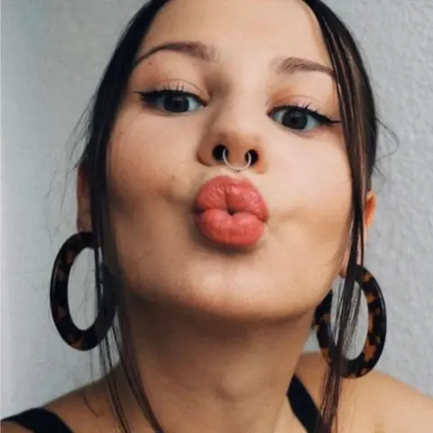 PA Media A young woman with brown hair and wearing bright red lipstick pouts at the camera. She is wearing a black vest top.