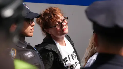 Reuters Susan Sarandon, whose ginger hair is tied up, wears glasses, a black hooded jacket and a white t-shirt, looks on as she is seen stood next to two police officers in New York City 
