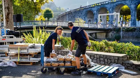 AFP via Getty Images Two workers, a man and a woman dressed in navy polo shirts and shorts, move cardboard boxes off a trolley in the Sanctuary of Our Lady of Lourdes.