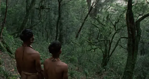 George Pimentel In this still, two men - father and son - are seen from behind look out over a dense, green forest.