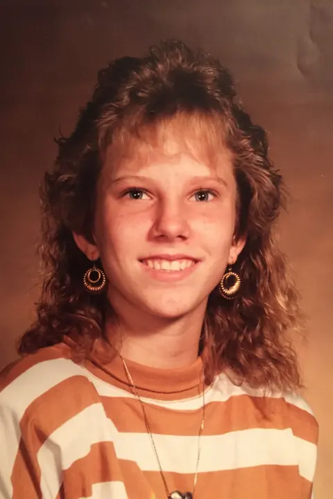 Family handout A portrait of Colleen Slemmer, taken in what looks like a photography studio with a backdrop. She is wearing a striped orange and white sweatshirt, gold drop earrings and is smiling at the camera. She has auburn hair and a fringe.