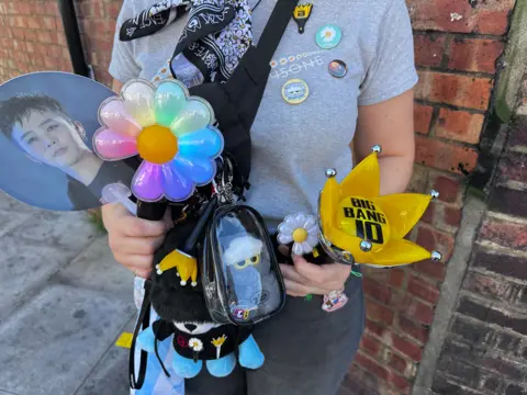 Person holding a collection of concert merchandise and light sticks in the foreground. Items include a flower-shaped light stick with pastel-coloured petals, a yellow crown-shaped light stick marked “BIG BANG 10”, a handheld fan featuring a portrait, and a small flower-shaped accessory. A black cross-body bag is decorated with badges and pins, and holds a clear pouch containing a blue character keyring. The person is standing beside a red brick wall, with a pavement visible to the left.