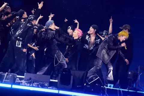 YG Entertainment A performer with bright yellow hair leans forward at the front of a stage, holding a handheld microphone. Around the performer, a group of performers and dancers in black outfits gather in a tight formation, with several raising their arms. Another performer with blue hair and a black outfit stands near the centre, while a performer with pink hair is visible beside them. They are illuminated by blue lighting against a dark background.