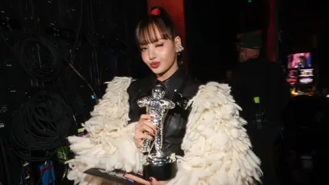 CBS via Getty Images Lisa stands backstage in a dimly lit production area, holding a silver astronaut-shaped trophy and a flat award plaque. She wears a black outfit with an oversized cream-colored textured coat featuring thick, looped fabric. One eye is closed in a posed expression.