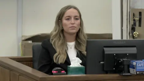 CBS Alleigha Botner testifies in court. She sits before a computer monitor and wears a black jacket, white shirt and shiny necklace and earrings