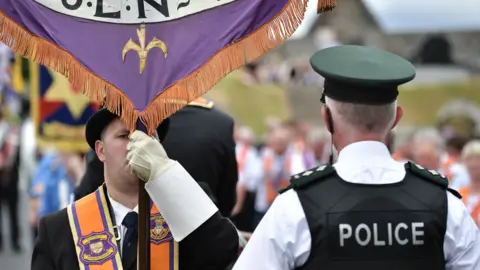 Getty Images A man is wearing an orange sash and a black bowler hat and holding a banner. A policeman has his back to the camera. He has a black vest on stating police in white lettering. He has a cap and a white shirt on. A parade is out of focus in the background. 