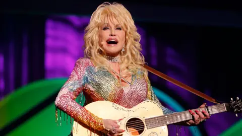 Getty Images dolly Parton performing on stage with a guitar in a multi coloured sequined jumpsuit. The staging is green and purple behind her