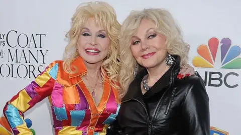 Getty Images Dolly Parton, wearing a bright and colourful dress, stands next to her sister Stella at a red carpet event in 2015 - who is wearing a black leather jacket and large jewellery. They are both smiling, with Dolly's arm around her sister's shoulders, as they are photographed.