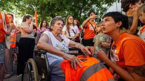 NurPhoto via Getty Images An elderly woman in a wheelchair is surrounded by protestors wearing orange in Madrid. 