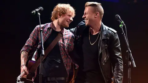 Getty Images Ed Sheeran and Macklemore on stage, in an embrace, during a performance, smiling at each other.