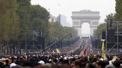Reuters Thousands of people can be seen lining Champs-Elysees Avenue. A sea of white hats and yellow tops line the street. There are several large scaffold structures with screens on and the Arc de Triomphe can be seen at the end of the street.