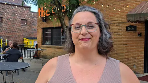 Brianna Beyrouti Brianna Beyrouti, wearing glasses and smiling, standing in front of what appears to be a cafe or restaurant, with two men sitting at outdoor tables behind her
