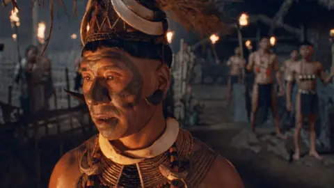 George Pimentel A man in traditional Konyak attire and facial tattoos stands in a village at night, illuminated by warm torchlight, with other figures visible in the background.