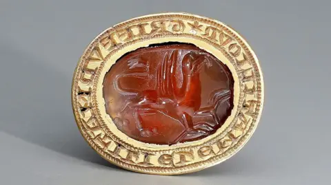 Noonans An oval of gold with an inscription engraved around its entire circumference. In the centre of the oval is a orange-red stone with a creature engraved into it. It appears to have a bird's head and a large body, a lion's hind legs and paws and bird's feet on its forelegs. 