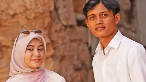 BBC Zia and Yusron smile at the camera - Zia is wearing a dusky pink hijab and has sunglasses on her head, Yusron is wearing an open neck white shirt 