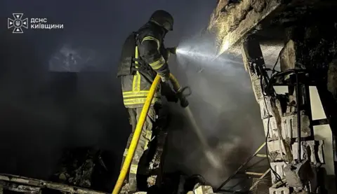 Kyiv State Emergency Service A firefighter wearing heavy duty black overalls with fluorescent green stripes holds a hosepipe next to a damaged buildings, which is at the scene of a Russian strike in the Kyiv region. 