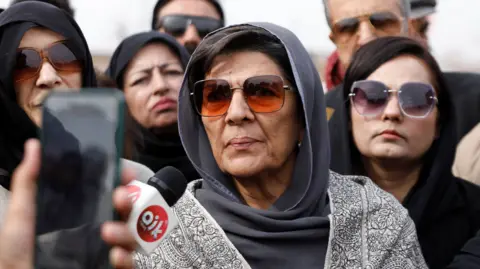 REUTERS/Salahuddin Aleema Khanum wears a grey head covering and sunglasses as she speaks into a news microphone. There are other women standing behind her.